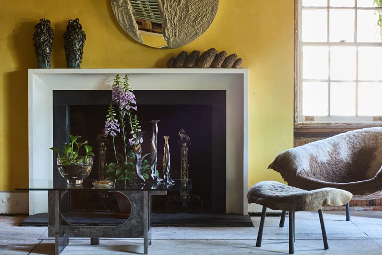 Stylish living room with a modern chair, glass table, art pieces, and fireplace against a yellow wall.
