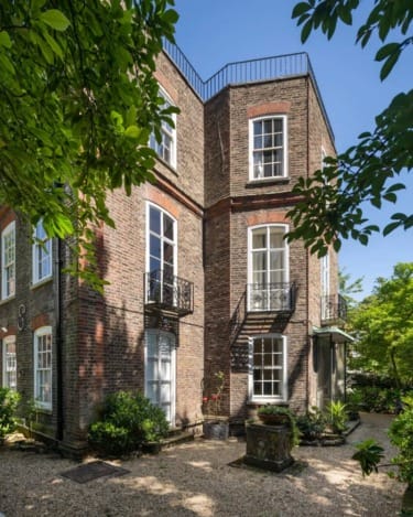 Three-story brick house with large windows, black railings, and surrounded by lush greenery on a sunny day.
