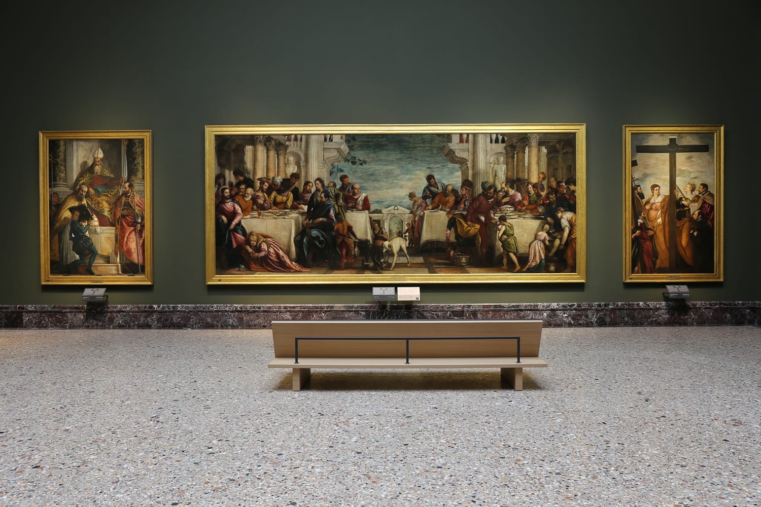 Art gallery with three large framed paintings on a green wall, viewed from behind a wooden bench in an empty room.