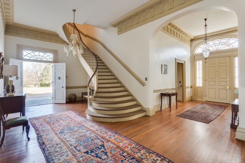 Spacious foyer with elegant curved staircase, wooden floors, chandeliers, and colorful Persian rugs.