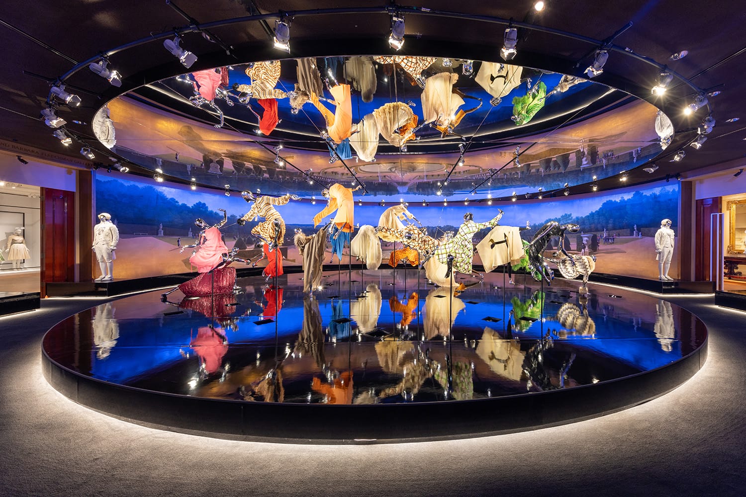 Exhibit featuring colorful, ornate garments displayed in a circular room with reflective floor and blue-lit landscape background.