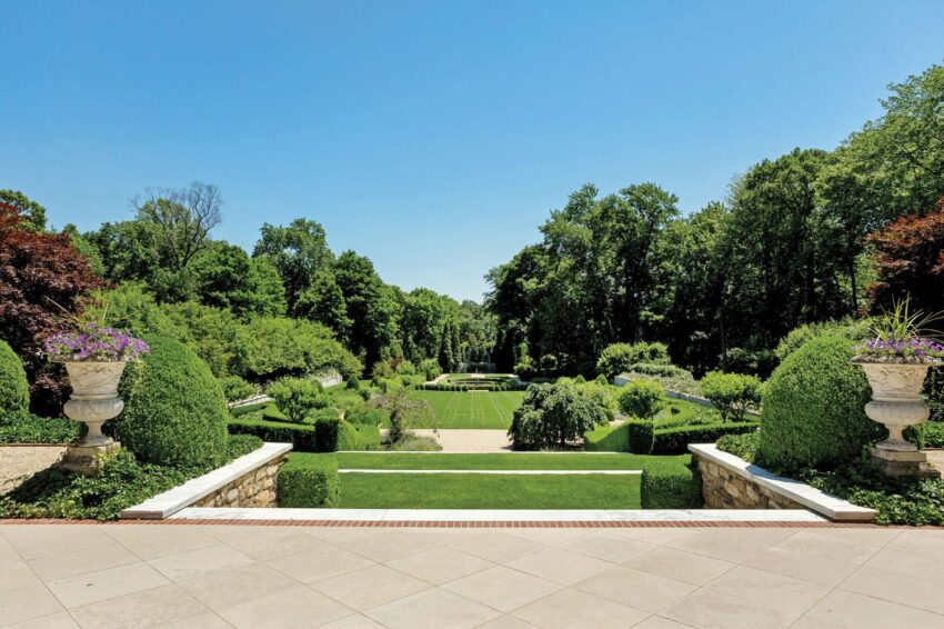 Elegant garden with manicured hedges, lush greenery, stone urns with flowers, under a clear blue sky.