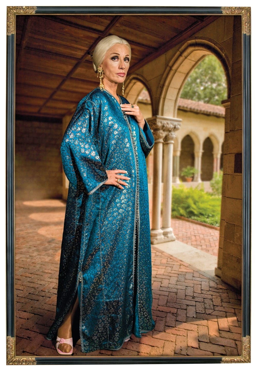 Woman in a blue patterned robe standing in a covered brick pathway with arches in the background.