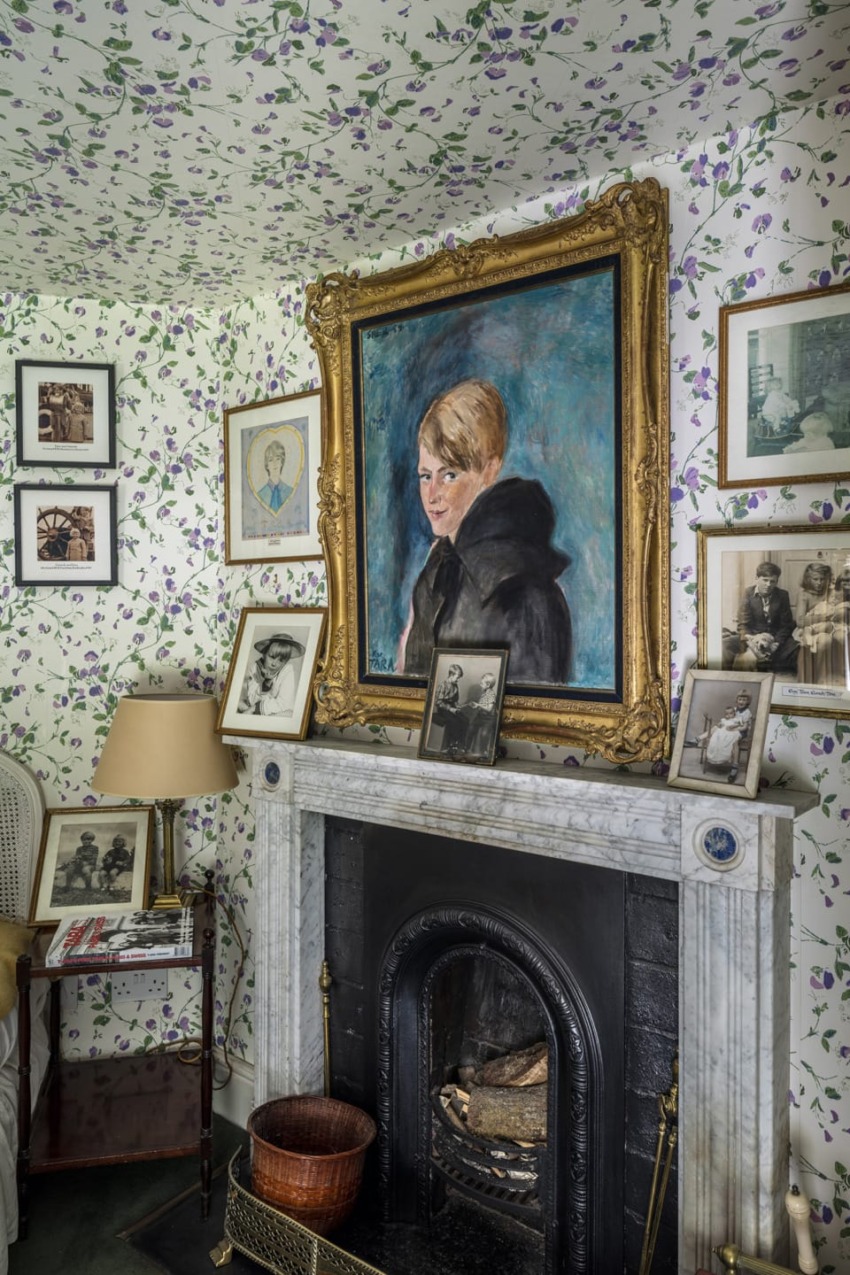 Cozy room with floral wallpaper, marble fireplace, and framed portrait above the mantle surrounded by vintage photographs.