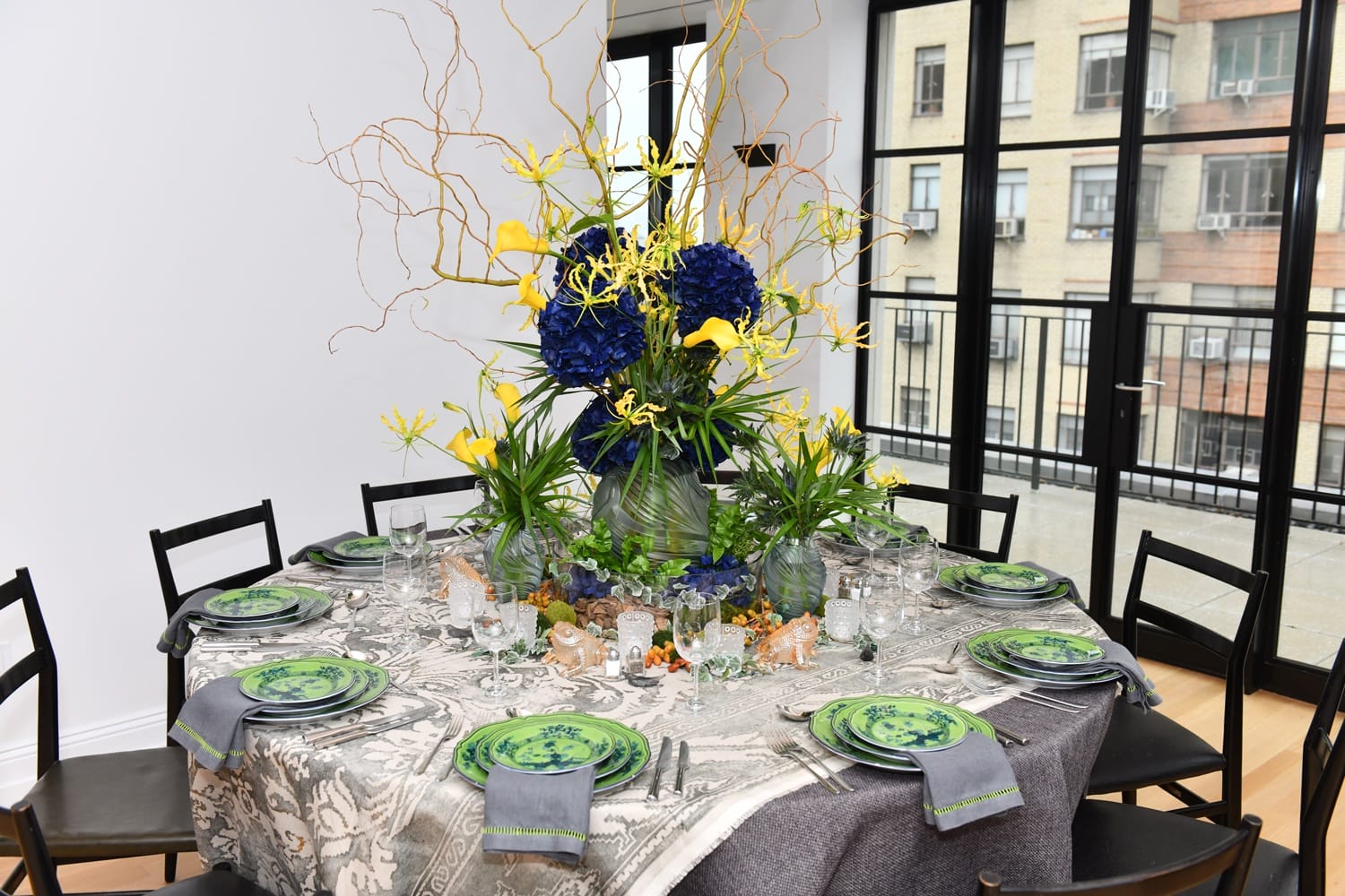 Elegant dining table with ornate green plates, floral centerpiece of blue and yellow flowers, surrounded by black chairs.