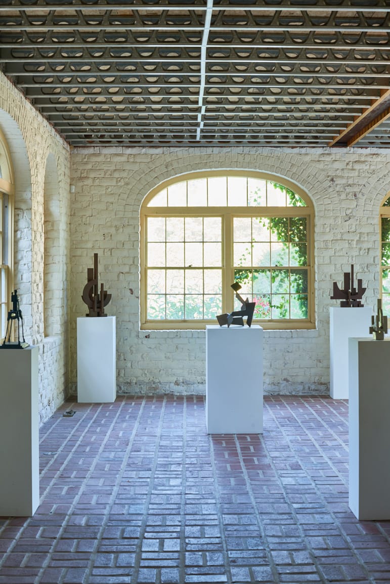 Art gallery with sculptures on white pedestals, brick floor, and arched windows letting in natural light.