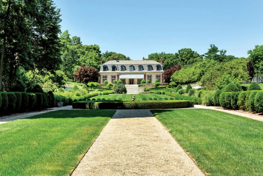 Elegant mansion with manicured gardens, lush green lawn, and a stone path leading to the entrance, surrounded by trees.