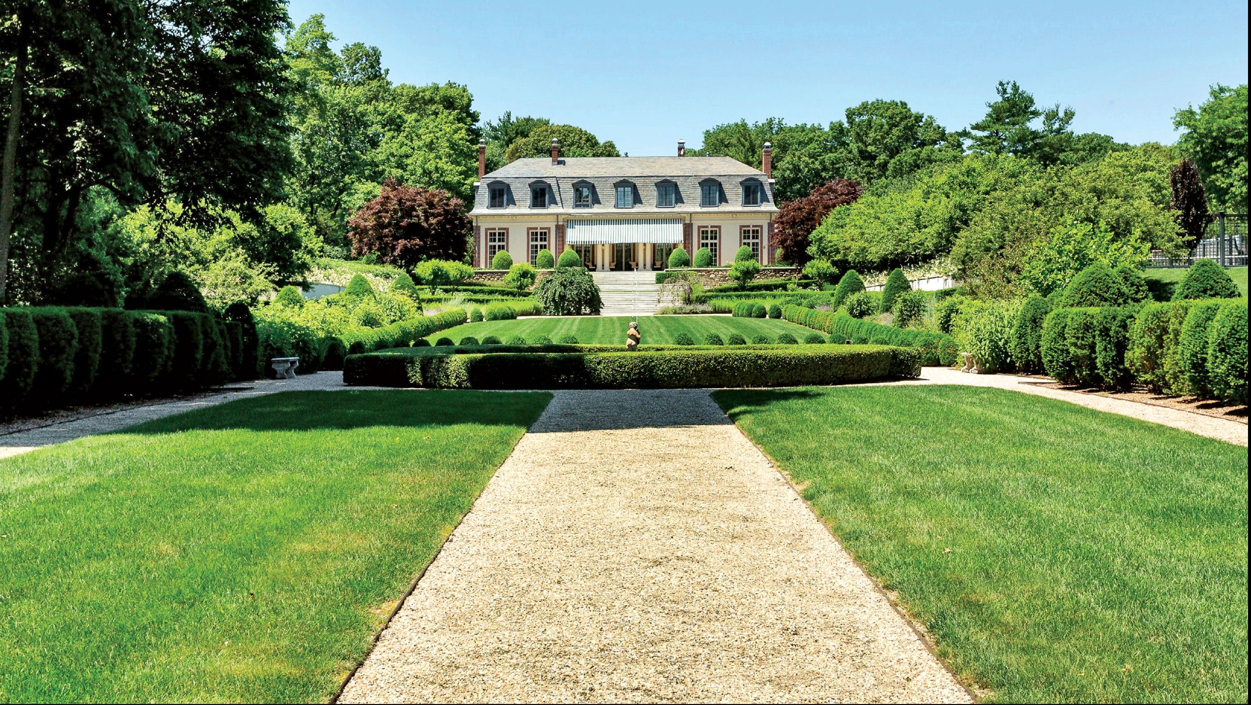 Elegant manor with manicured gardens, trimmed hedges, and a gravel pathway leading to the house, framed by lush greenery.