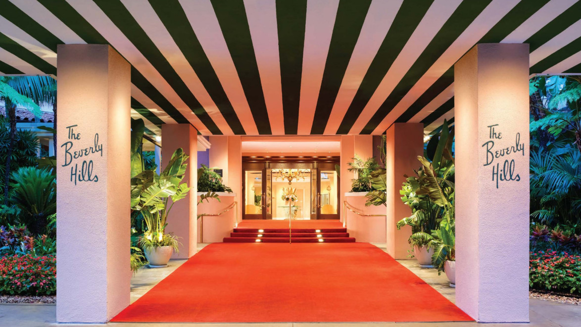 Entrance with red carpet lined by greenery and striped ceiling, leading to a glass door, at a luxury hotel.