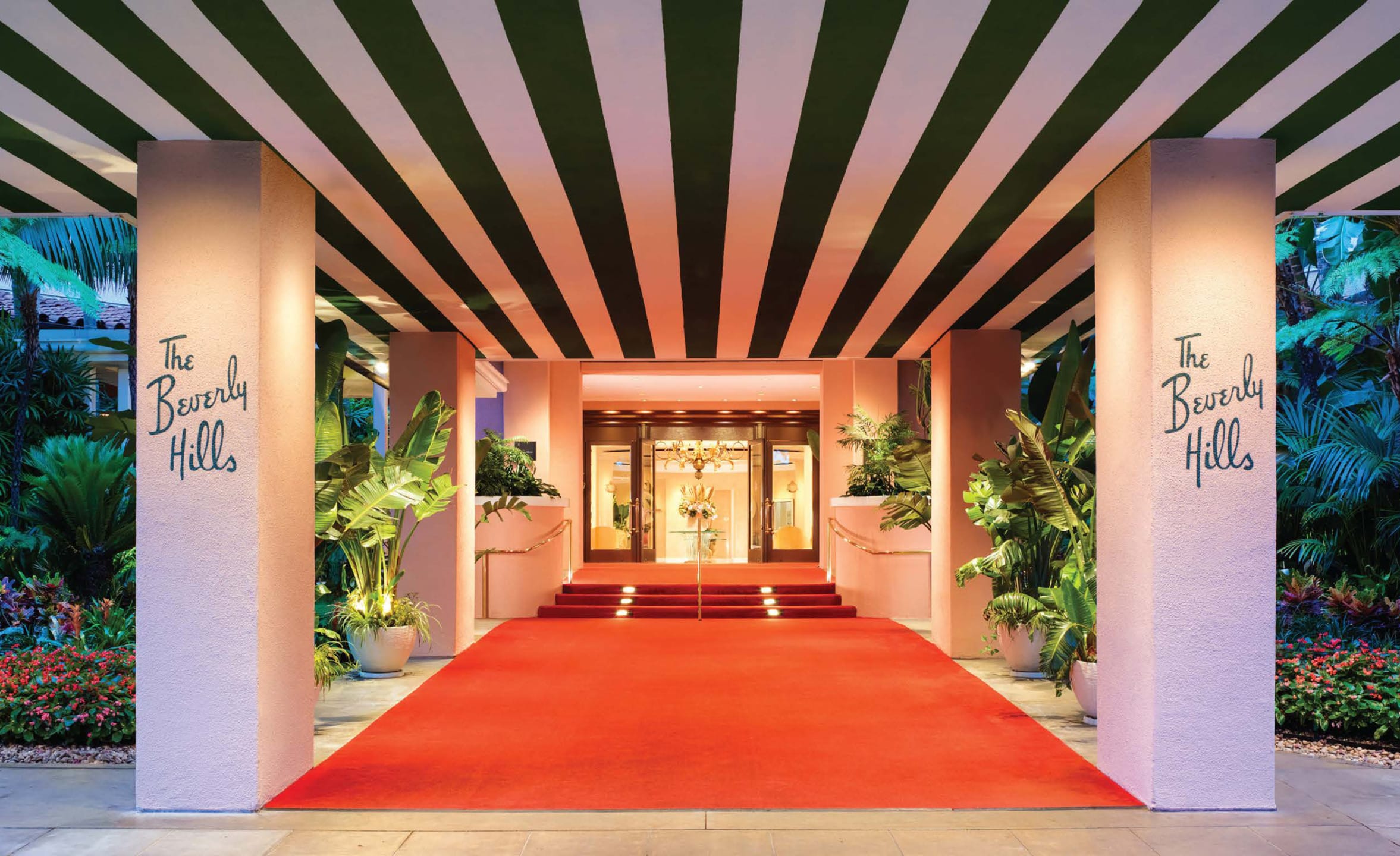 Entrance with red carpet lined by greenery and striped ceiling, leading to a glass door, at a luxury hotel.