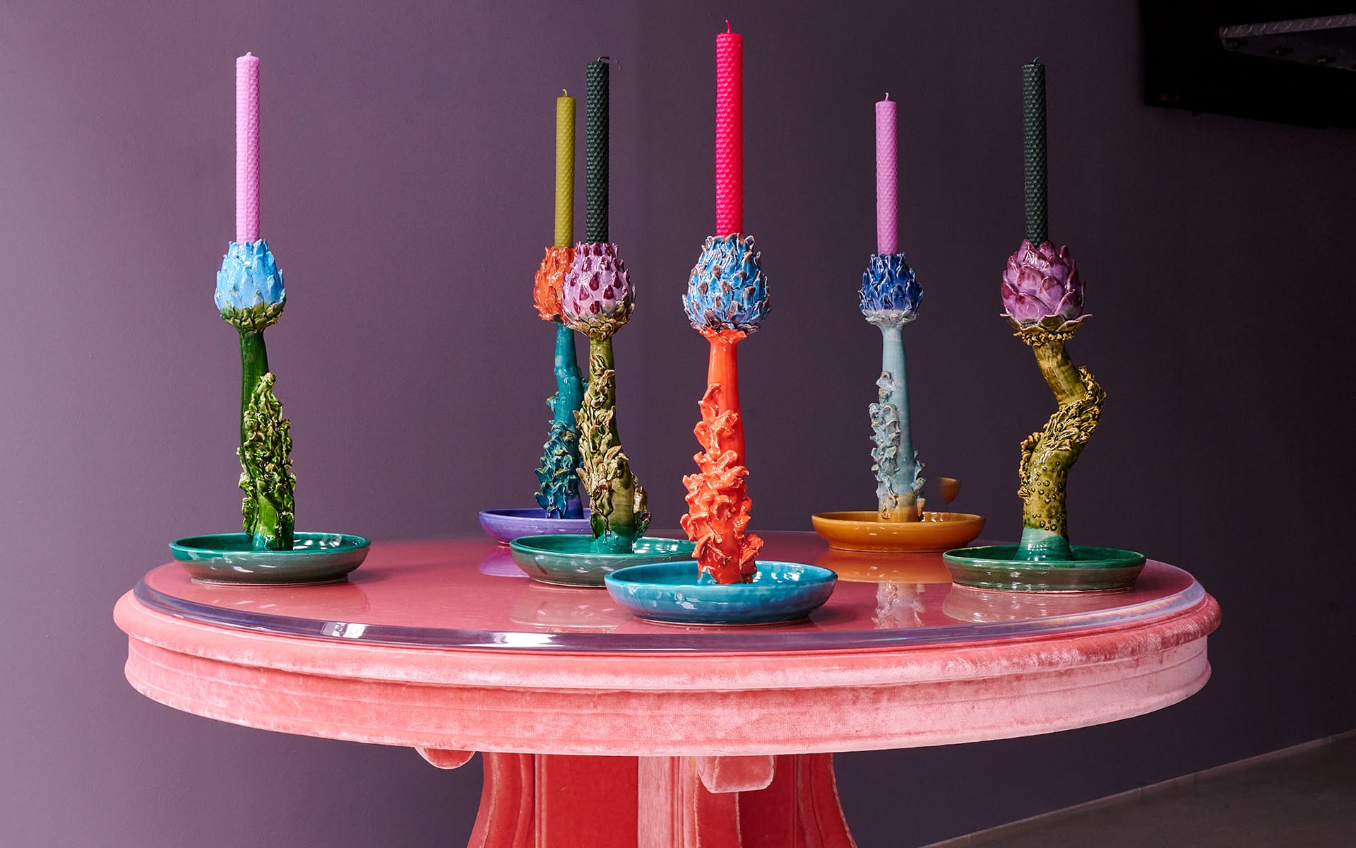 Colorful sculptural candleholders with tall, textured candles on a round pink table against a purple wall.