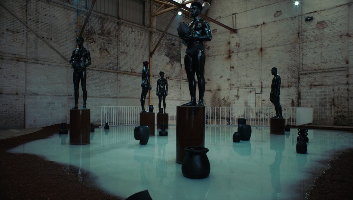 Art installation featuring dark humanoid sculptures on pedestals in an industrial space with a reflective white floor.