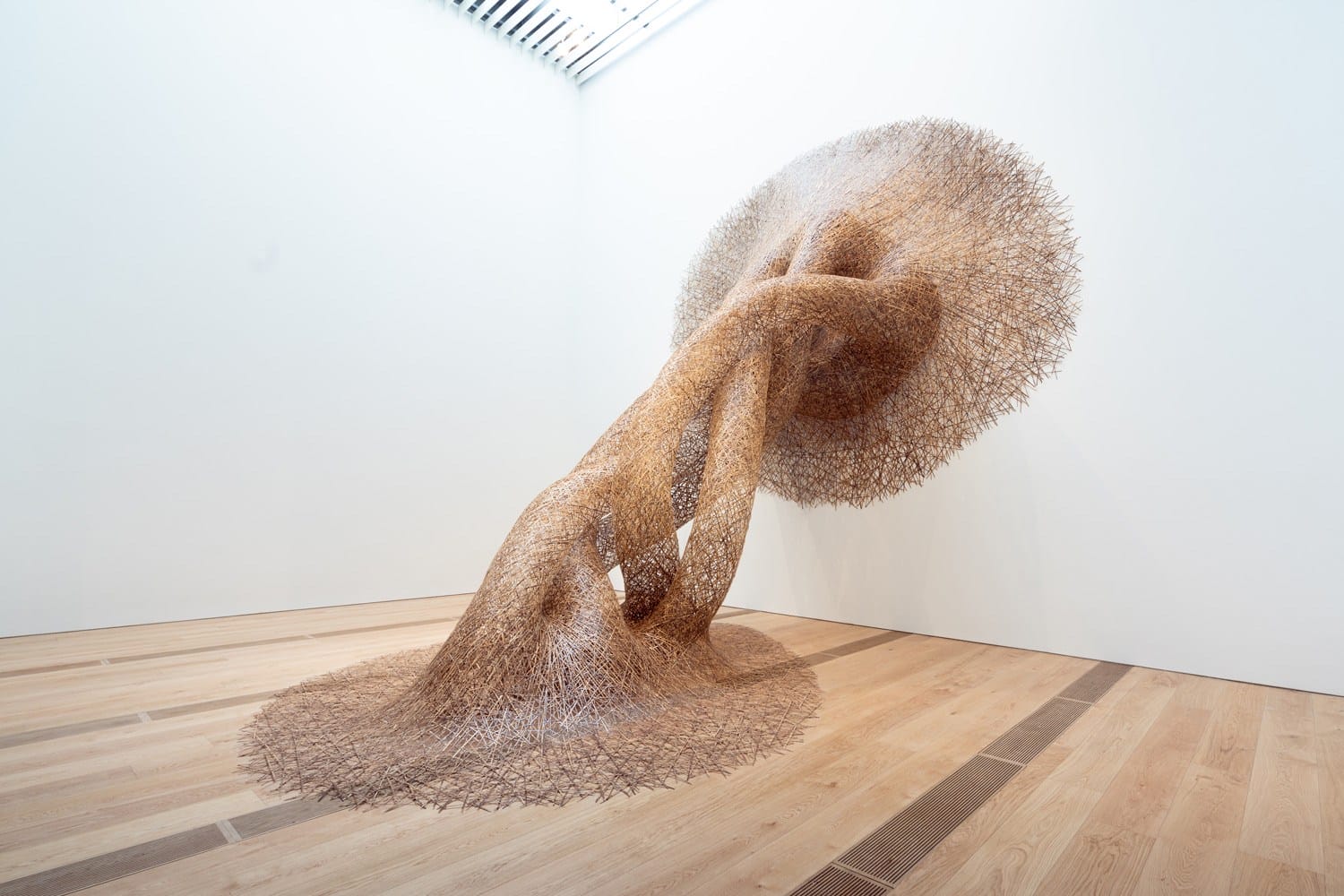 Intricate woven sculpture resembling a large mushroom with twisting stems, displayed in a modern gallery with wooden floors.