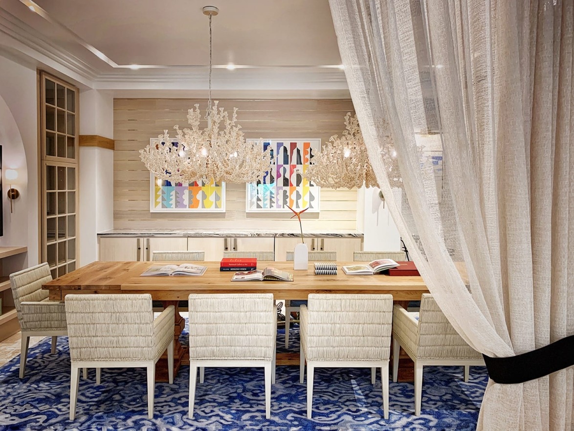 Elegant dining room with a long wooden table, white chairs, artistic paintings, and ornate chandeliers behind sheer curtains.