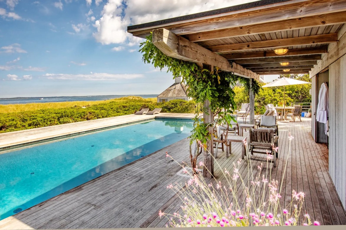 Outdoor patio with wooden pergola and seating beside a turquoise pool, overlooking a scenic landscape and bright blue sky.