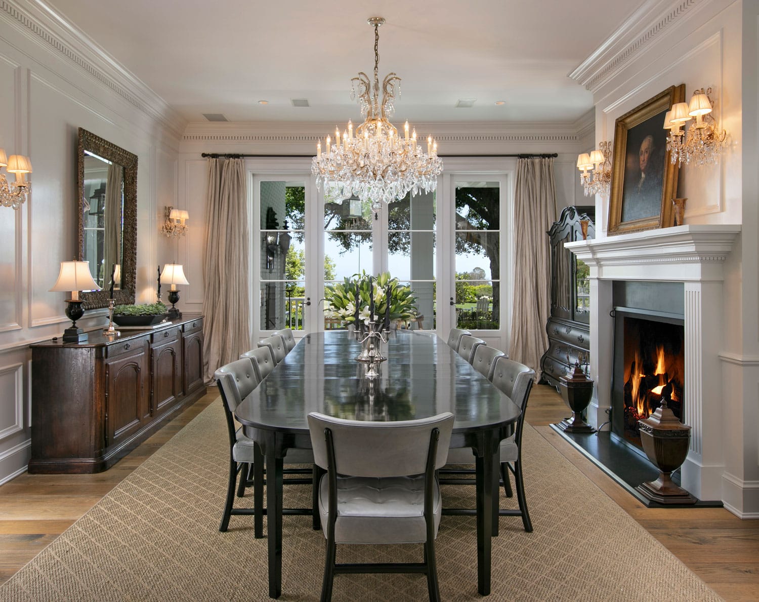 Elegant dining room with chandelier, large table, fireplace, and decorative artwork above the mantel in bright, airy setting.