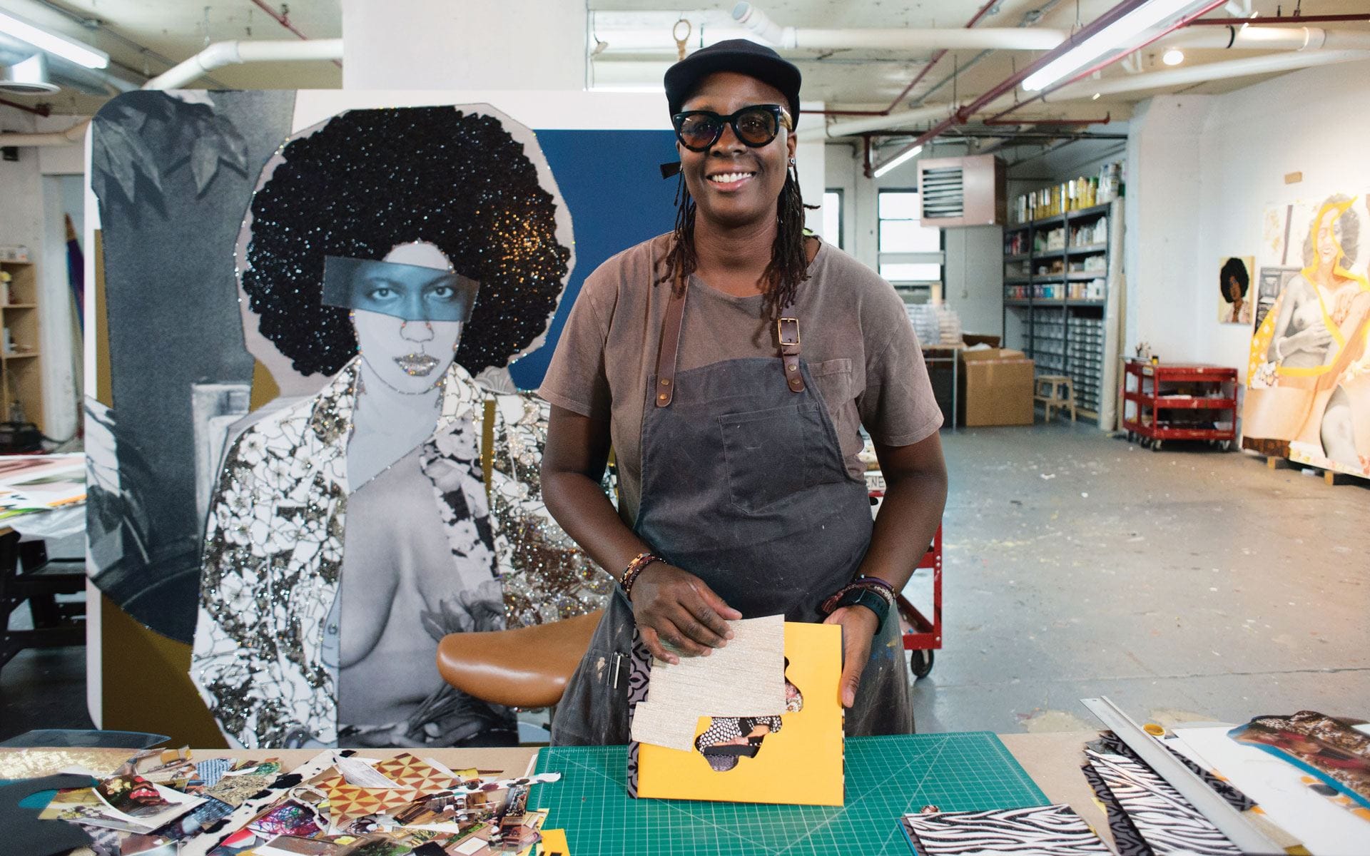 Artist smiling in studio, working on a collage with colorful materials, large artwork in the background.