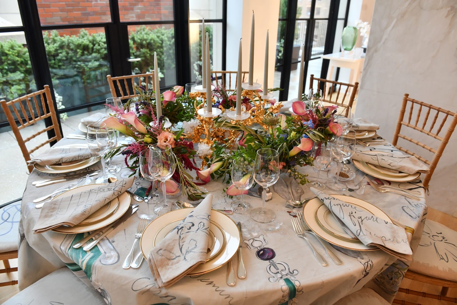 Elegant table setting with floral centerpiece, candles, glassware, and plates arranged on a patterned tablecloth.