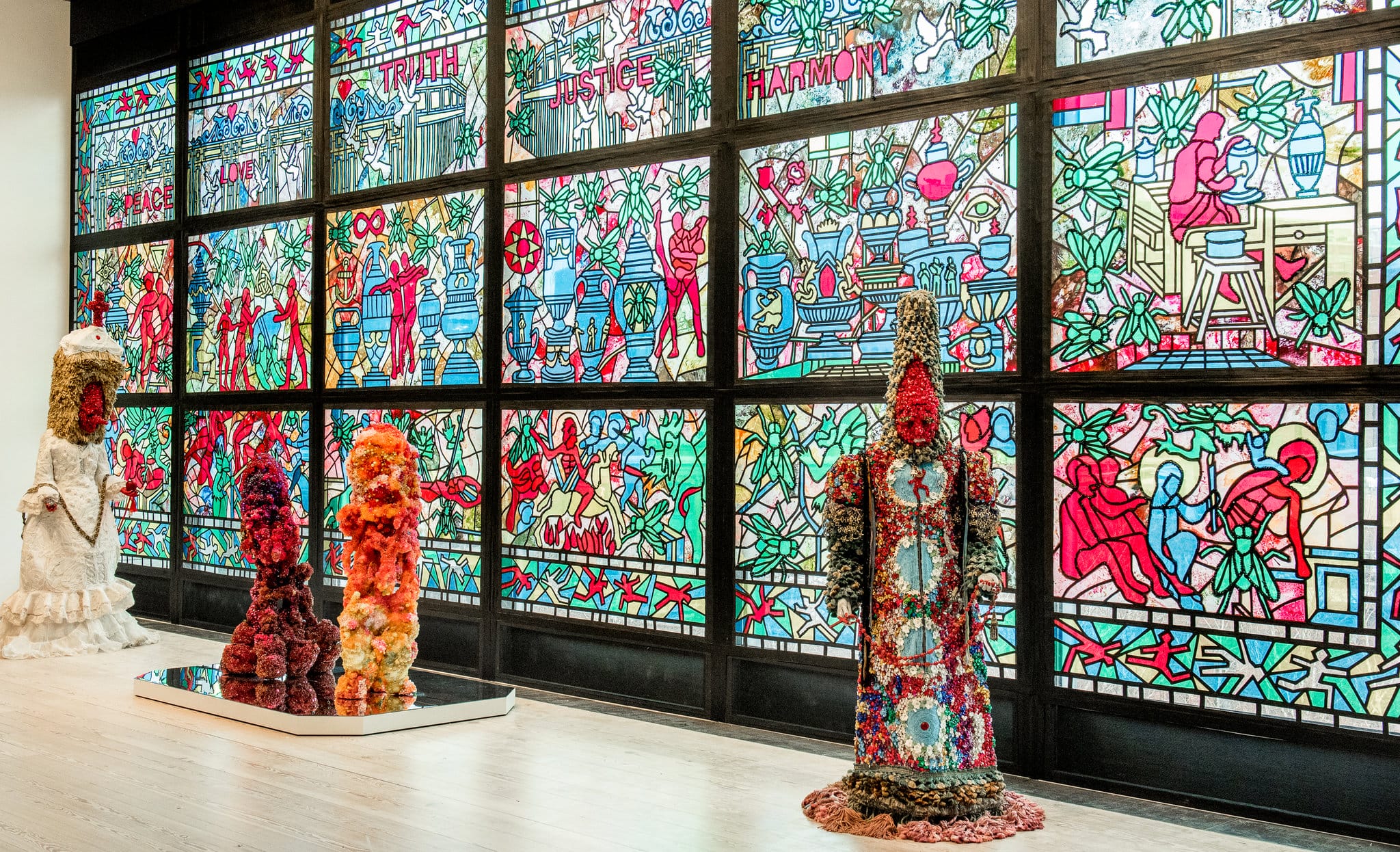 Colorful stained glass wall with abstract patterns and four unique textile sculptures displayed in a modern art gallery.