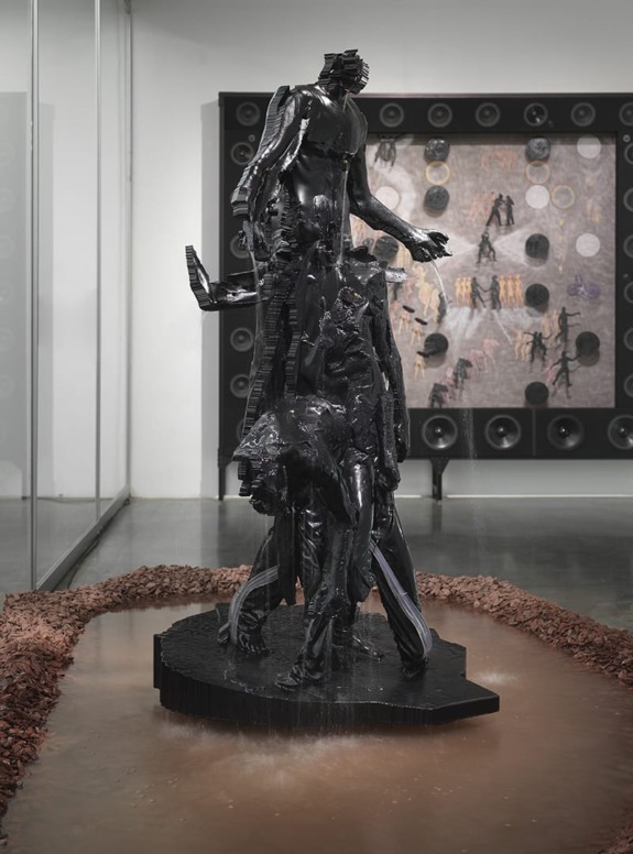 Abstract black sculpture with multiple figures in a gallery setting, surrounded by a water feature and artwork on the wall.