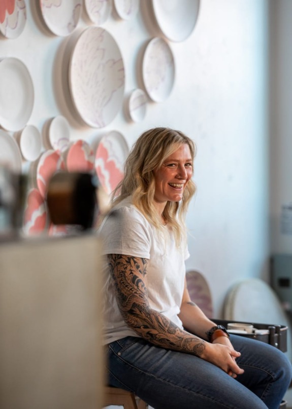 Person with tattoos sitting and smiling in a room with artistic plates on the wall.