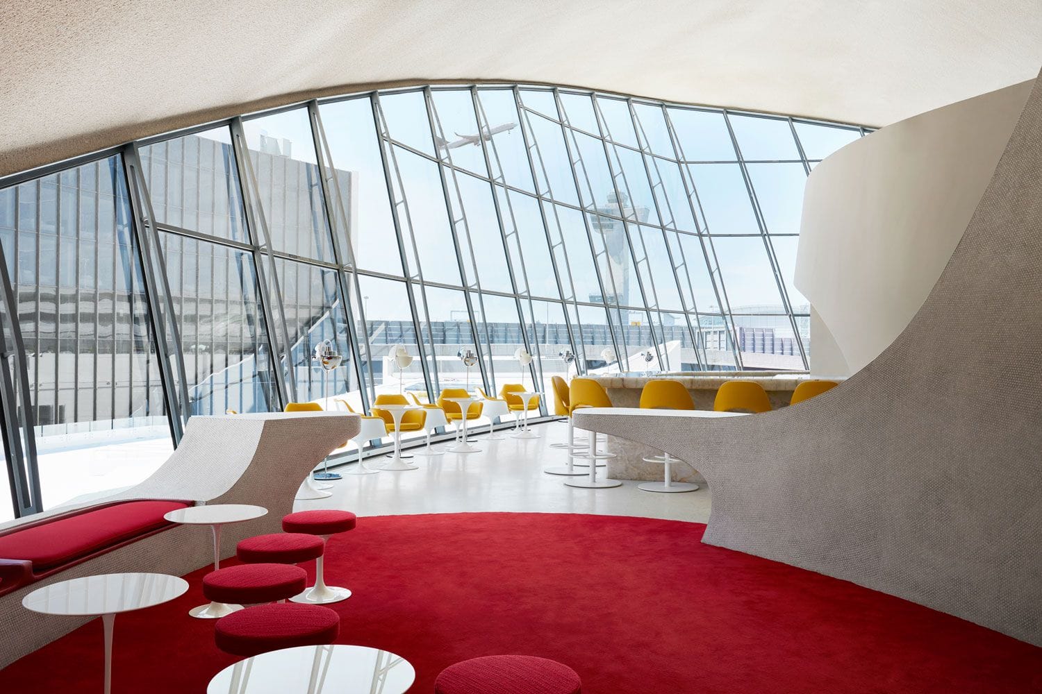 Modern lounge with red carpet, yellow chairs, floor-to-ceiling windows, and view of tarmac through curved glass wall.