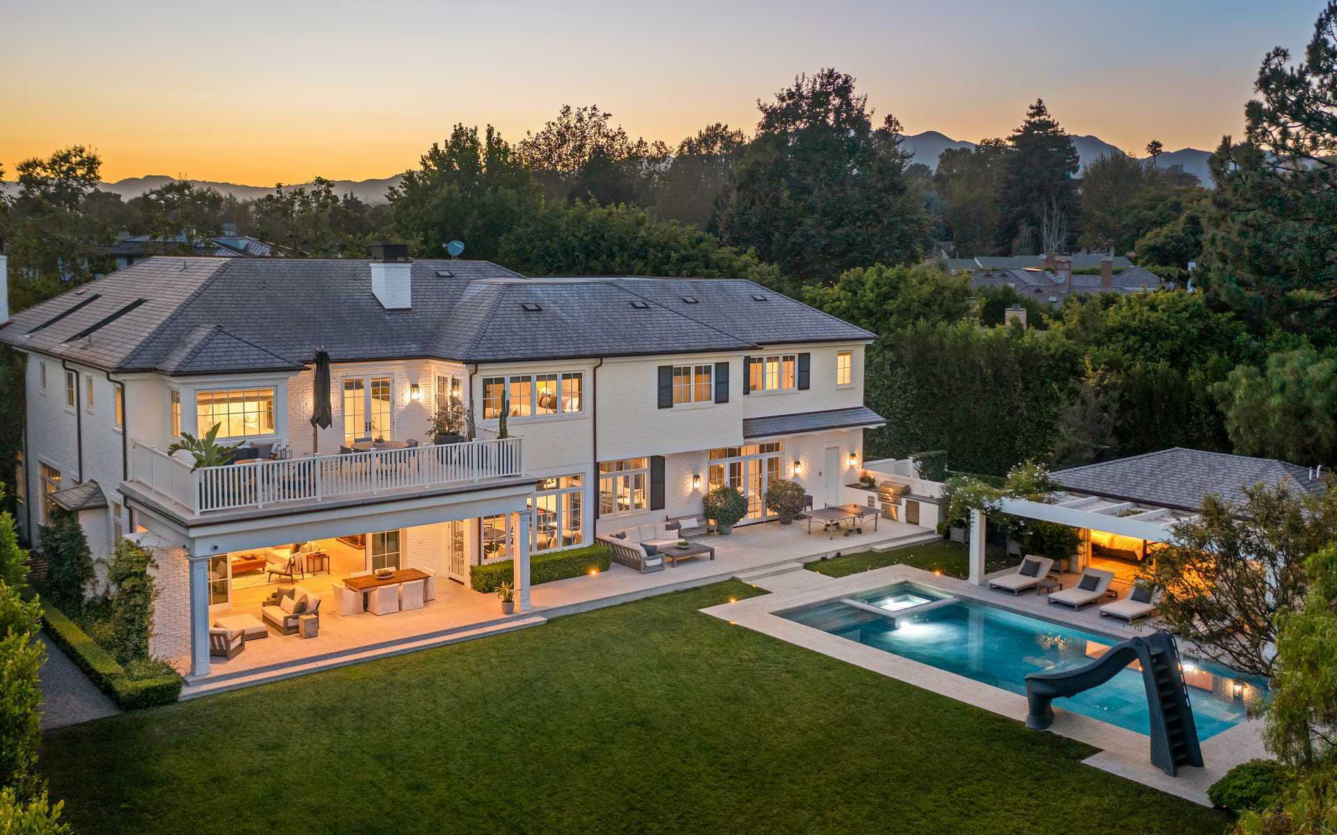 Luxurious two-story house with a pool, surrounded by lush greenery and outdoor seating, during sunset.