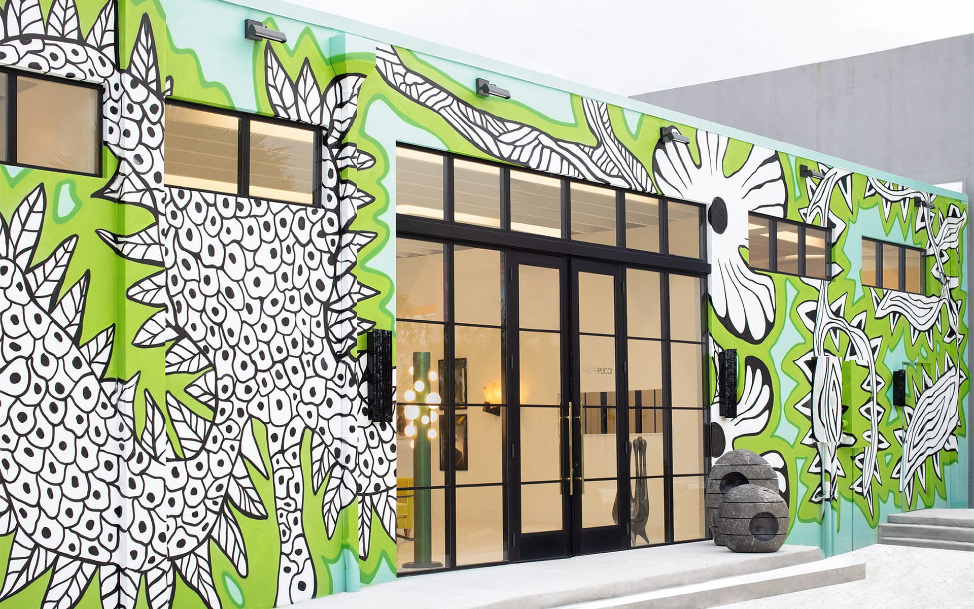 Colorful building exterior with green and black abstract mural, featuring plants and geometric patterns surrounding glass doors.