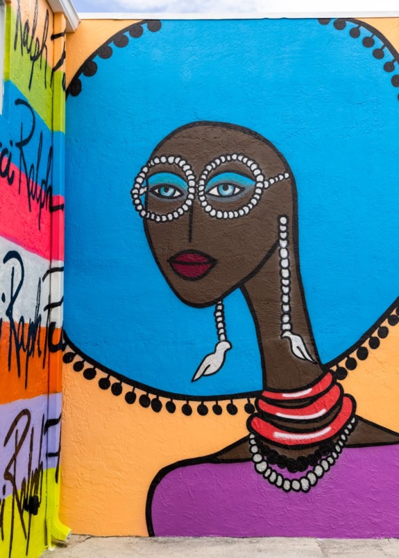 Colorful mural of a woman with a large blue hat, beaded earrings, and glasses, against a vibrant graffiti background.