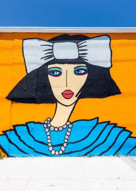 Colorful mural of a woman with a large white bow, pearl necklace, and stylized black hair against an orange wall.
