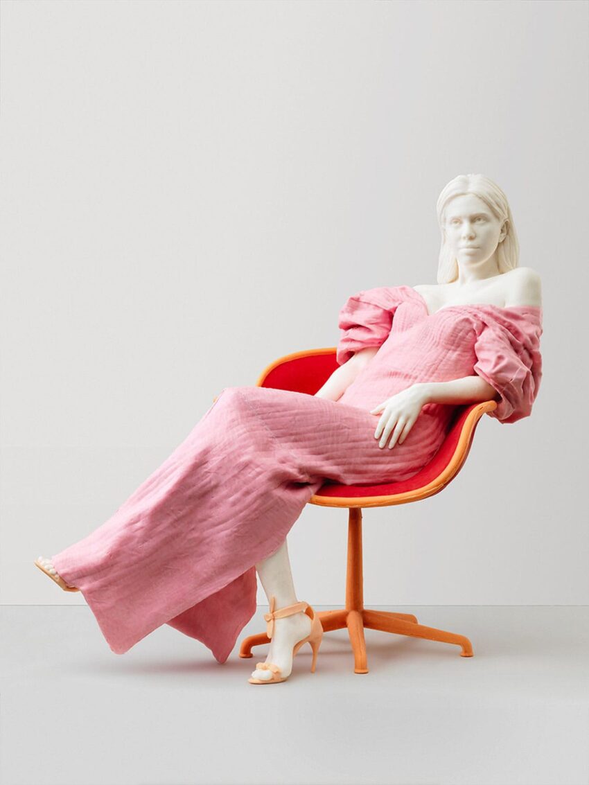 Sculpture of a woman in a pink dress seated on an orange chair against a neutral background.