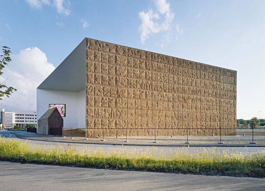 Modern building with a textured facade, large overhang, and a small annex on a sunny day with a blue sky background.