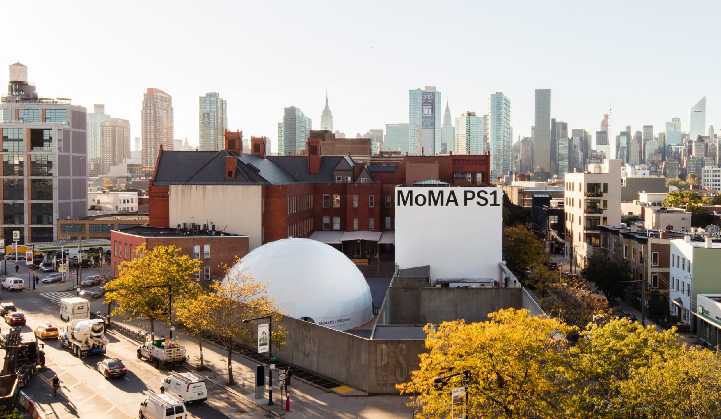 View of MoMA PS1 art center with surrounding urban skyline and trees in autumn colors, located in Long Island City, New York.