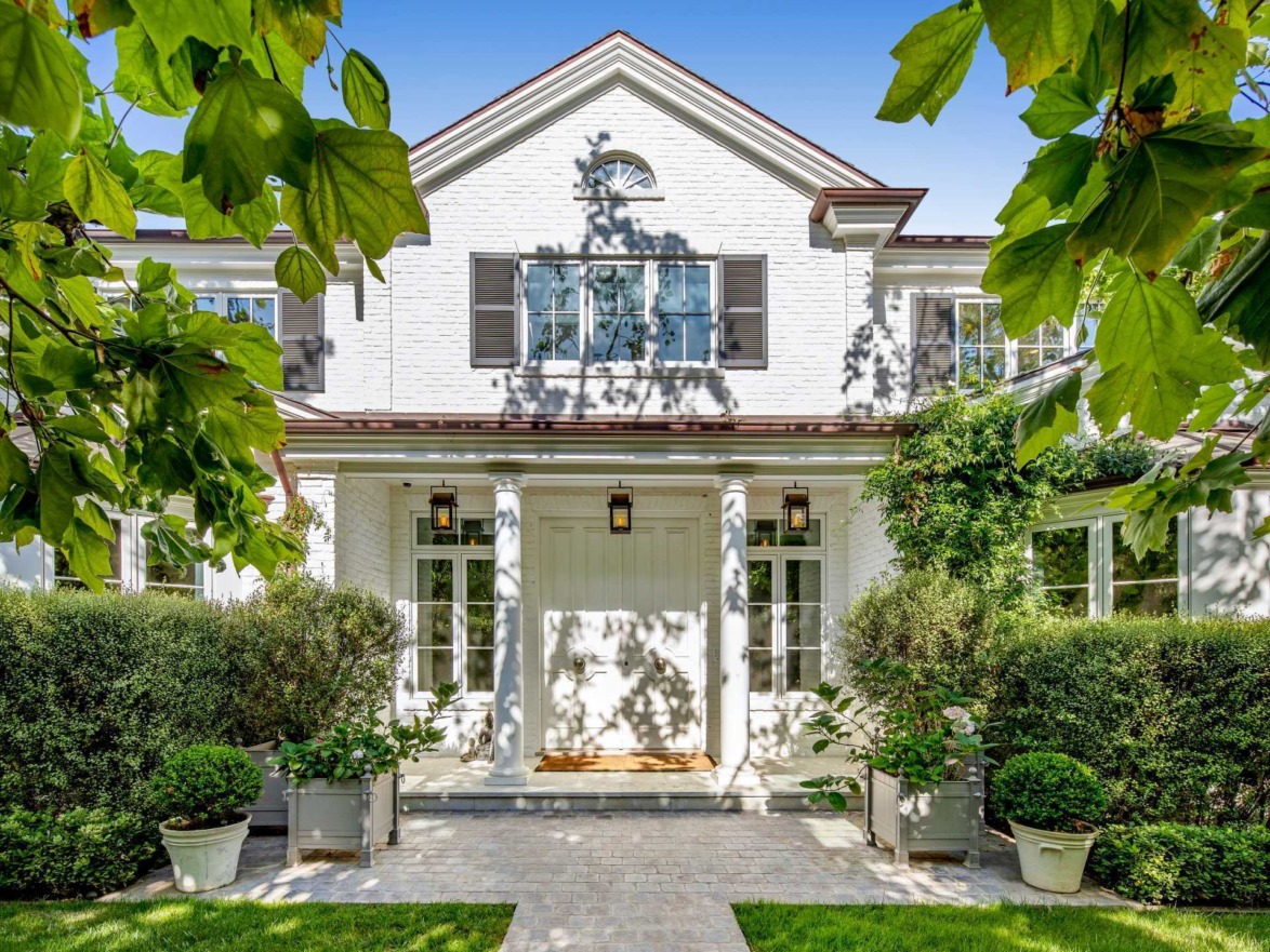 Front view of a two-story white brick house with green plants and trees, featuring large windows and a central entrance.