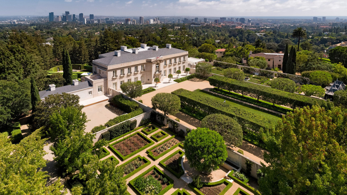Aerial view of a large mansion surrounded by formal gardens and city skyline in the distance under a cloudy sky.