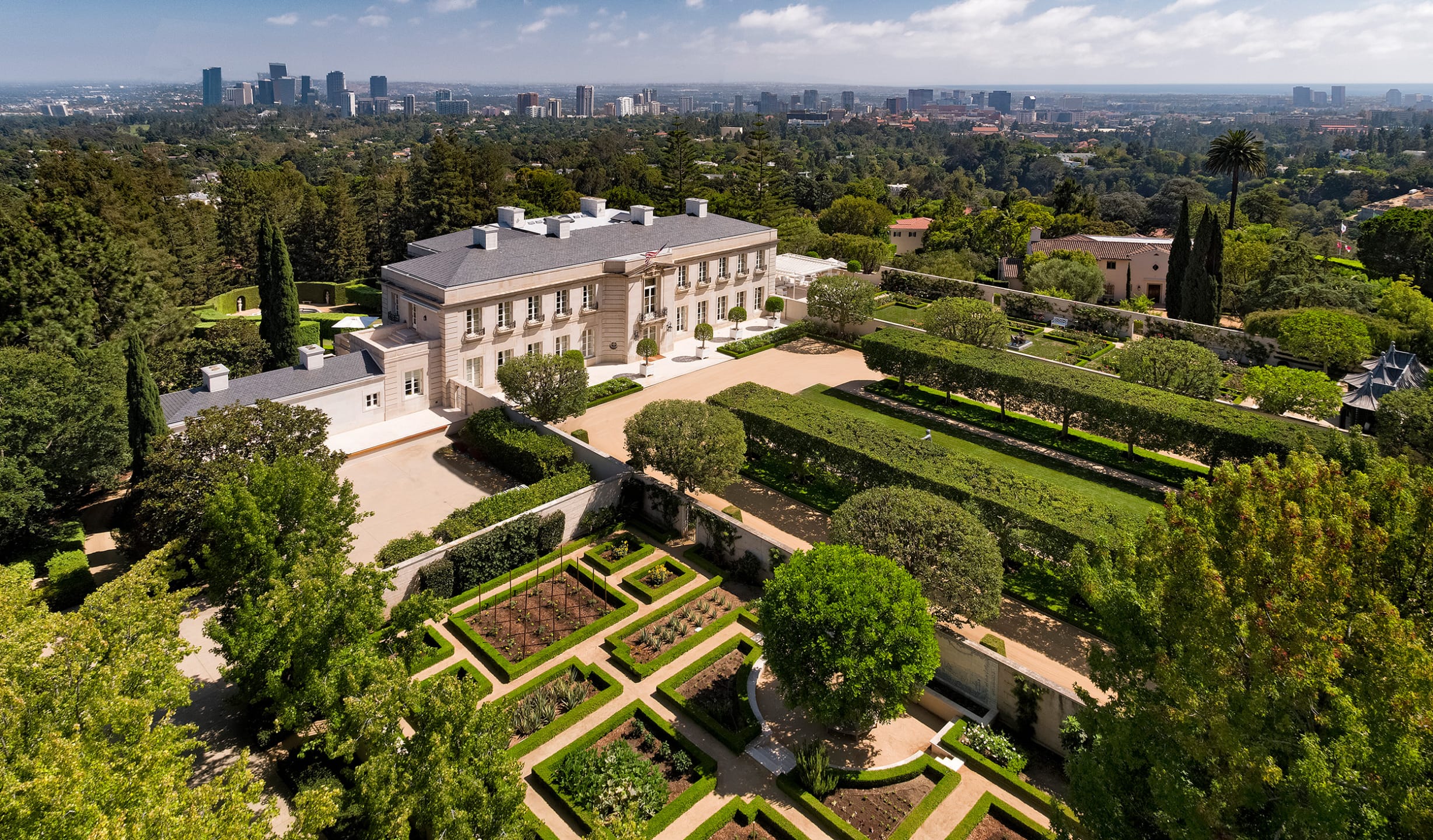 Aerial view of a large mansion surrounded by formal gardens and city skyline in the distance under a cloudy sky.