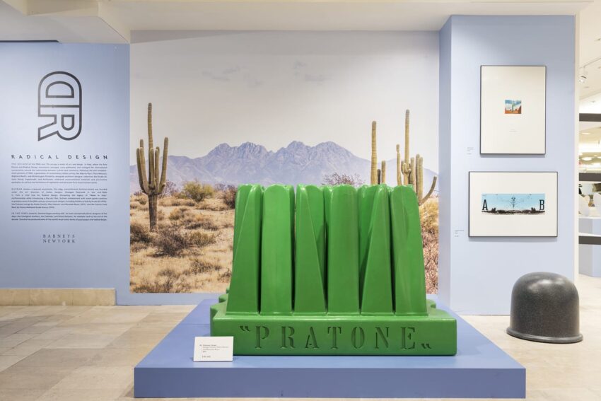 Green Pratone chair sculpture resembling oversized grass, displayed in a modern art gallery with a desert mural background.