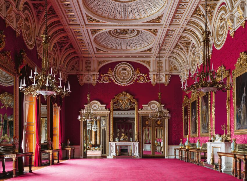 Opulent room with red walls, high ornate ceiling, chandeliers, gold detailing, large mirrors, and classic paintings.