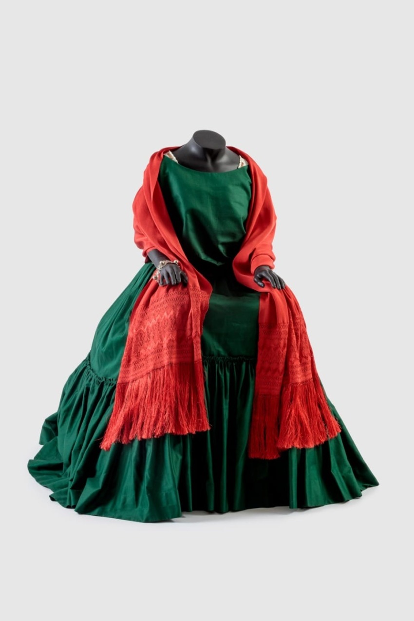 Victorian-style gown in green with red shawl draped over mannequin, no head or arms, on light background.