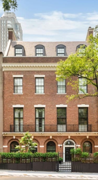 Brick townhouse with large windows, black iron fences, and green trees in an urban setting.
