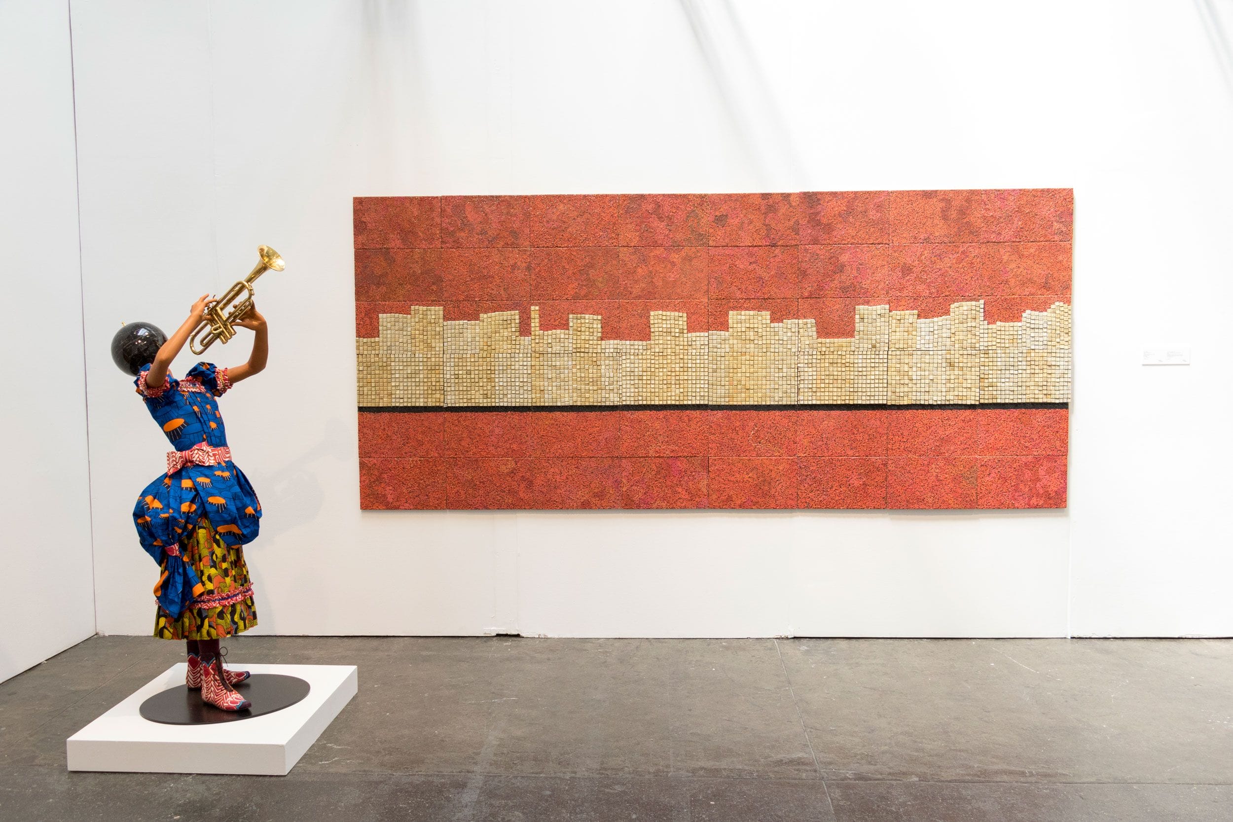 Sculpture of a woman playing trumpet beside a wall artwork with red and beige abstract cityscape pattern in a gallery setting.