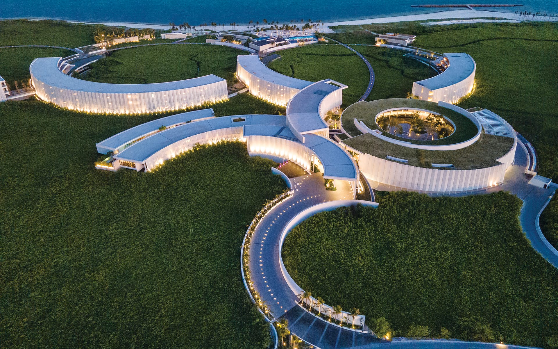 Aerial view of a modern, architecturally unique hotel with circular design surrounded by greenery and a coastal backdrop.