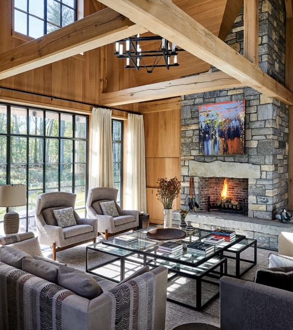 Rustic living room with wooden beams, stone fireplace, cozy chairs, and large windows letting in natural light.