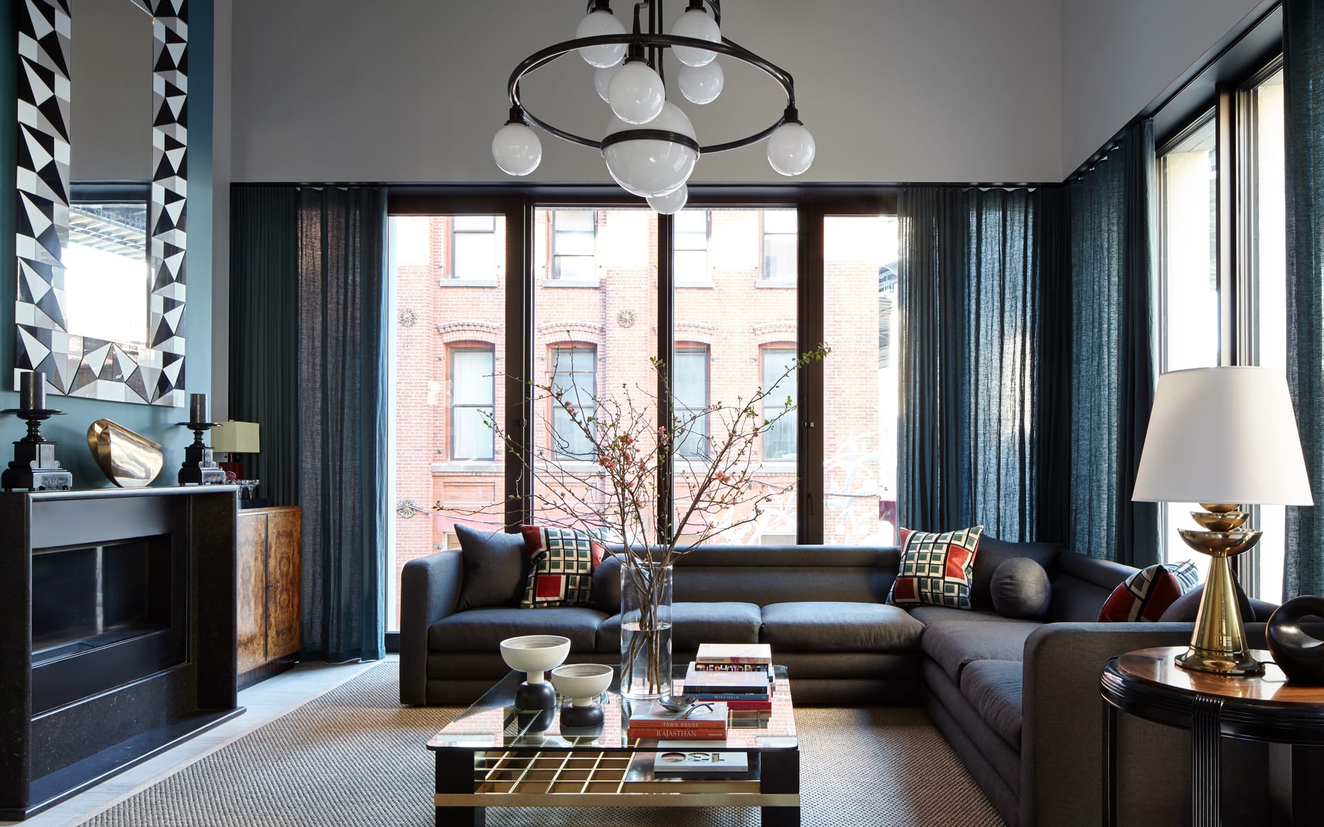 Modern living room with large windows, dark gray sectional sofa, stylish chandelier, and decorative accents.