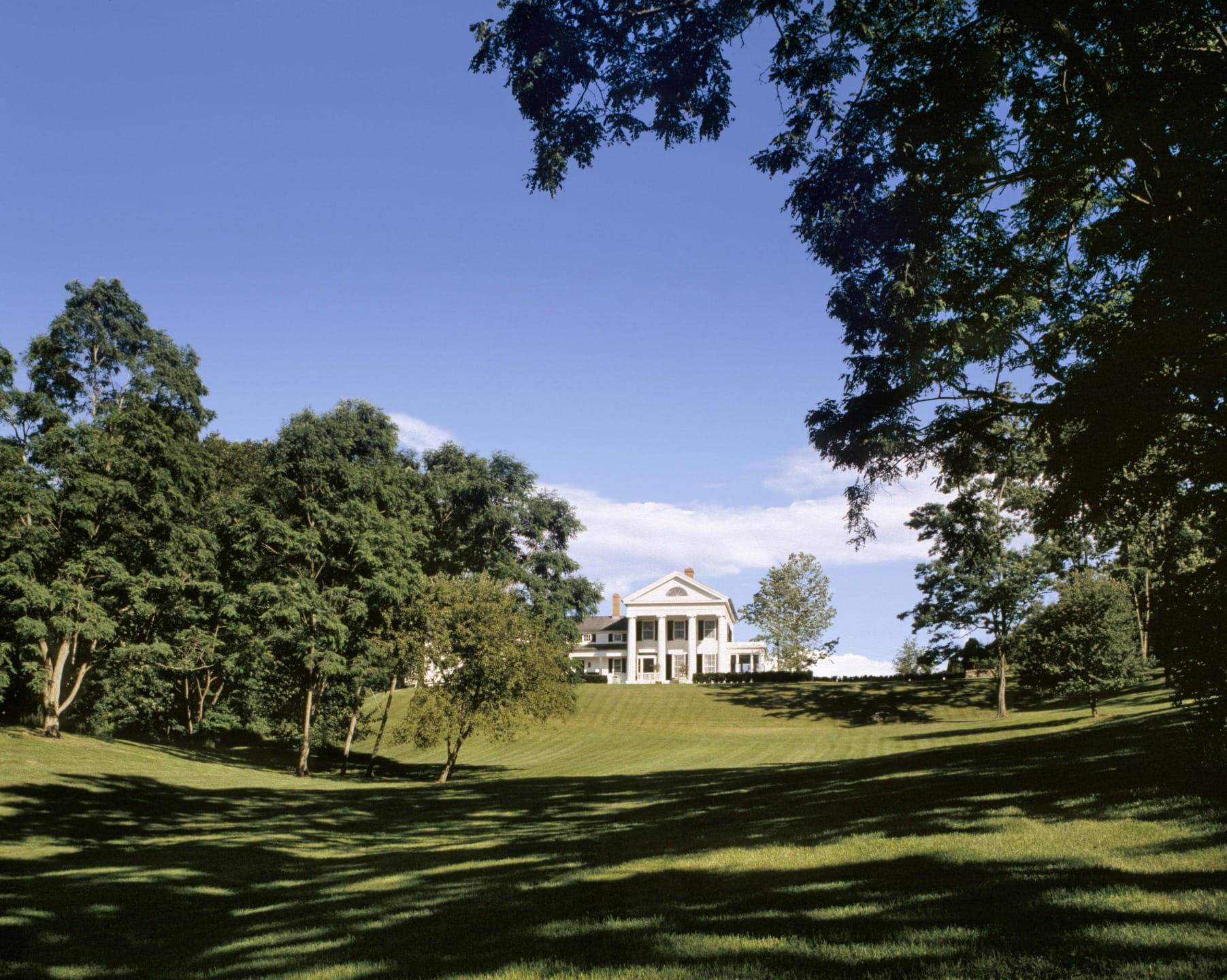 Large white mansion surrounded by lush green lawn and trees under a clear blue sky.