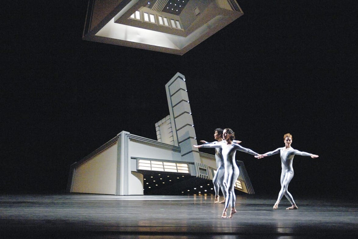 Dancers in metallic suits perform on stage with a futuristic set design in the background.