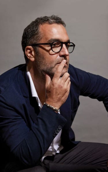 Man in dark blazer, wearing glasses, sitting and thoughtfully resting his hand on his chin against a gray background.