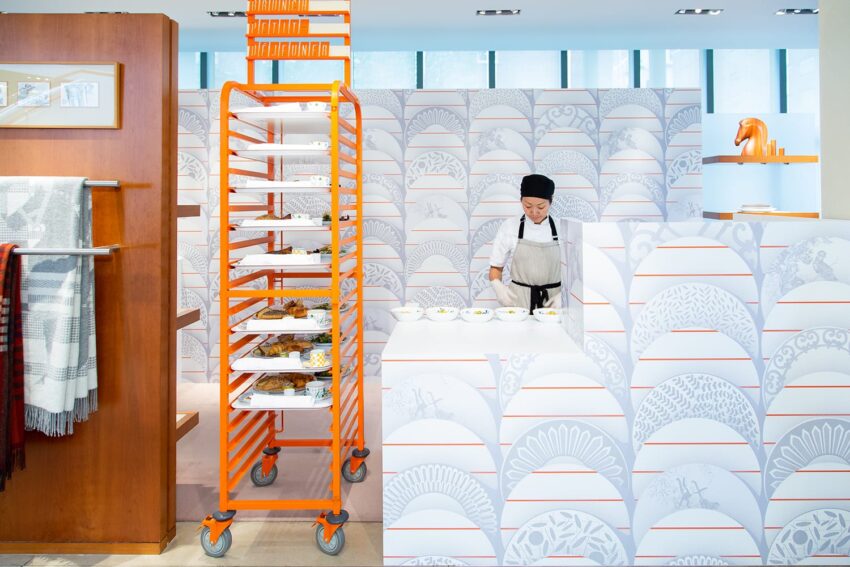 Chef preparing dishes at a stylish counter with patterned wall and orange rack holding trays in a modern setting.