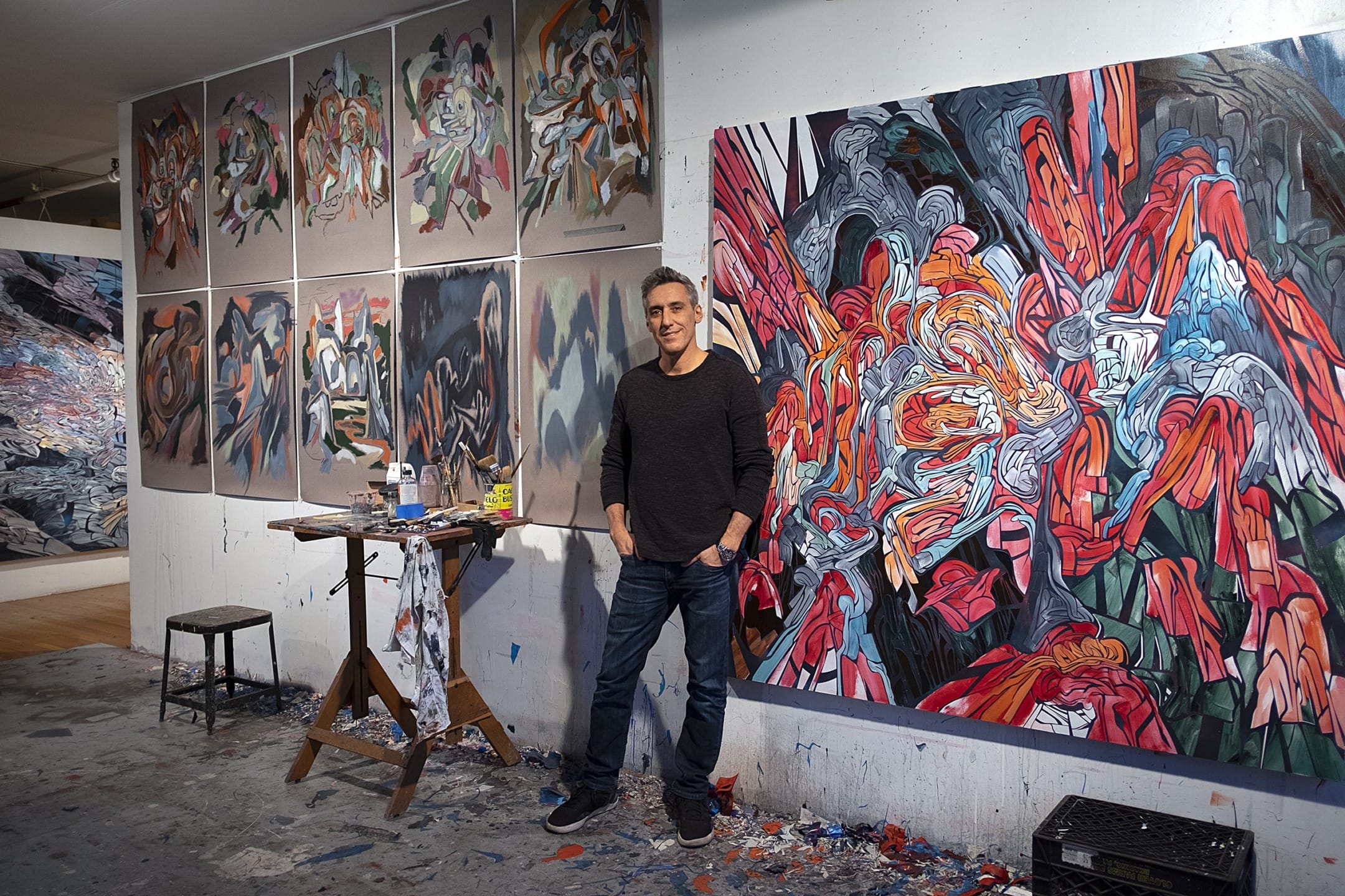 Artist standing in studio surrounded by colorful abstract paintings on walls and easel.
