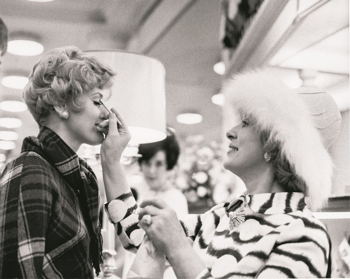 Two women in stylish vintage outfits, one applying makeup to the other, in a department store setting.